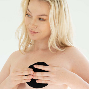 Woman looking into a mirror applying the black shorty pre-cut shape tape on her left breast. 