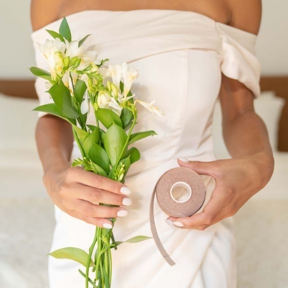 Woman is wearing a white strapless wedding dress while wearing boob tape and nipple covers underneath. In her right hand she is holding a bouquet of white flowers. In her left hand she is holding a roll of beige boob tape.
