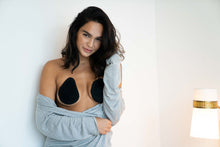Load image into Gallery viewer, Woman leaning against a white wall with her left arm touching her hair. Woman is wearing a grey cardigan draped over her arms. Woman is wearing black shorty cut pre cut tear drop shape tape on both breasts.
