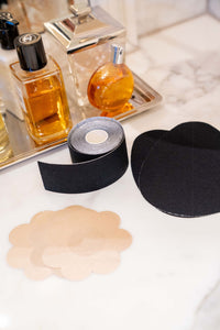 Nipple covers, a black boob tape roll and black classic cut tear drop pre-cut shape tape on a dresser with a tray of perfume in the background. 